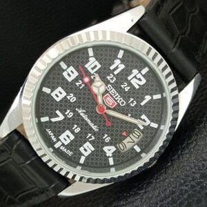 May include: A Seiko 5 automatic wristwatch featuring a black textured dial with white Arabic numerals, a 24-hour inner scale, and a date window. The watch has a silver-toned fluted bezel, luminous hands with a red second hand, and a black leather strap.