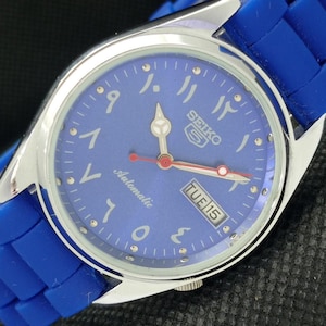 May include: A Seiko 5 automatic wristwatch with a round silver-tone stainless steel case and a vibrant blue silicone strap. The blue dial features Eastern Arabic numerals, silver-tone hands, a red second hand, and a day-date window displaying TUE 15.