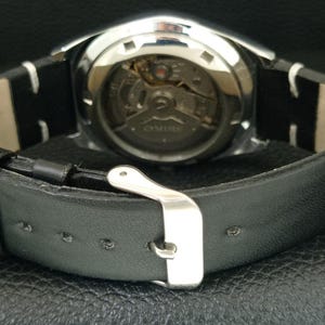 May include: Close-up of a silver-toned watch with a black leather band. The watch face is visible, showcasing the intricate internal mechanisms. The band features a silver buckle and white stitching.