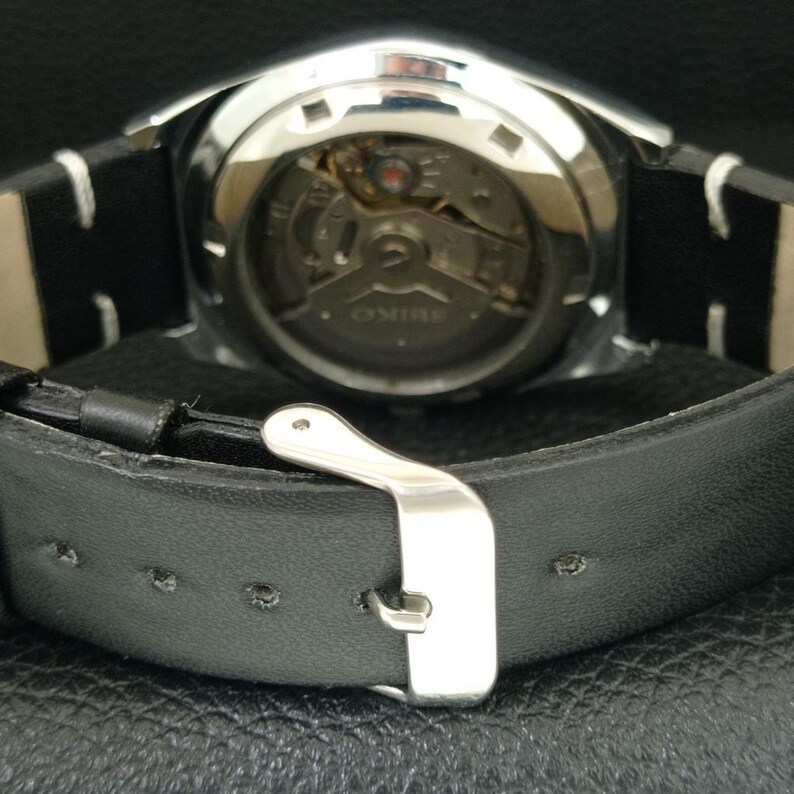 May include: Close-up of a silver-toned watch with a black leather band. The watch face is visible, showcasing the intricate internal mechanisms. The band features a silver buckle and white stitching.