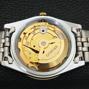 Peut inclure: Gros plan d'une montre Seiko avec un bo&icirc;tier et un bracelet argent&eacute;s. Le cadran transparent r&eacute;v&egrave;le le mouvement m&eacute;canique dor&eacute; complexe. Le mot "SEIKO" est visible sur le mouvement.