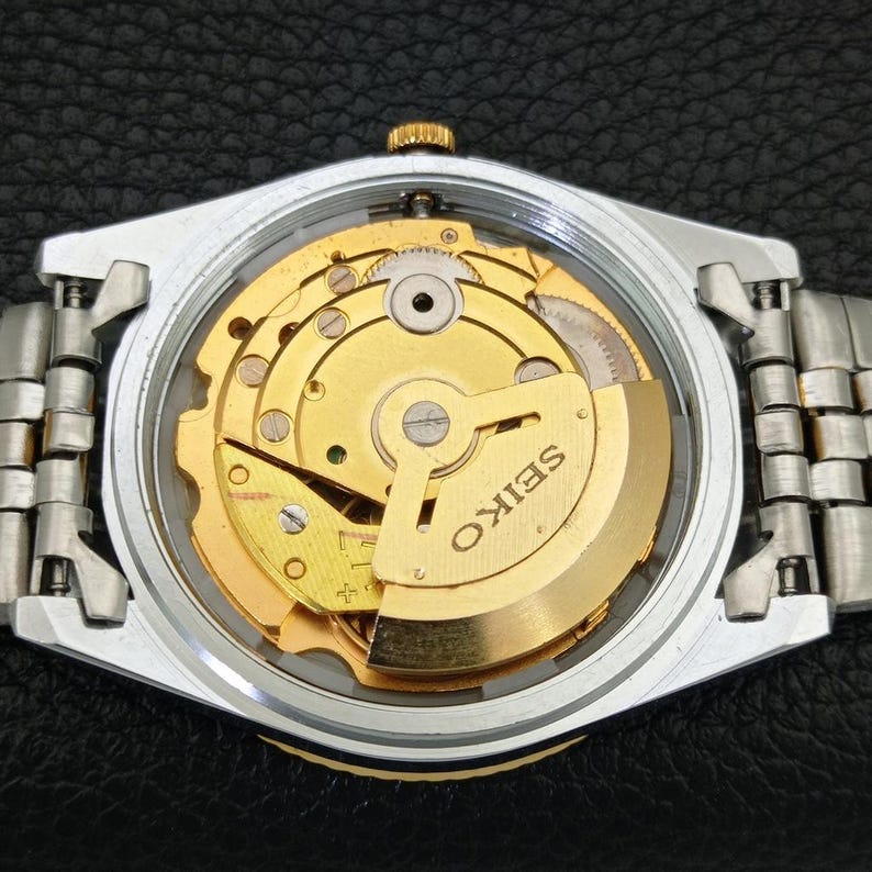 Peut inclure: Gros plan d'une montre Seiko avec un bo&icirc;tier et un bracelet argent&eacute;s. Le cadran transparent r&eacute;v&egrave;le le mouvement m&eacute;canique dor&eacute; complexe. Le mot "SEIKO" est visible sur le mouvement.