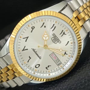 Op de afbeelding: Seiko 5 automatisch polshorloge met een zilveren wijzerplaat en goudkleurige lunette en wijzers. Het horloge heeft Arabische cijfers, een dag-datumweergave en een tweekleurige roestvrijstalen en gouden armband.