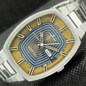 Puede incluir: Un reloj de pulsera automático Fortis Eden Roc plateado con una esfera cuadrada y una esfera azul y dorada. El reloj tiene una ventana de fecha en la posición de las 3 en punto y las palabras "Fortis Eden Roc Automatic" están impresas en la esfera.
