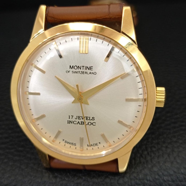 K&ouml;nnte beinhalten: Eine goldfarbene Armbanduhr mit rundem silbernem Zifferblatt, beschriftet mit Montine of Switzerland, 17 Jewels, Incabloc und Swiss Made. Die Uhr hat goldfarbene Zeiger und Indizes sowie ein braunes Lederarmband mit strukturierter Oberfl&auml;che.