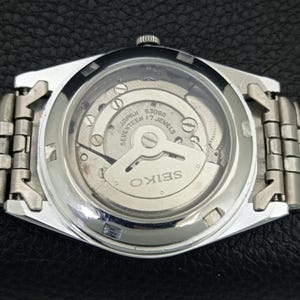 May include: A silver-toned Seiko watch with a transparent back, showcasing the intricate inner workings of the mechanism. The watch face displays the text "JAPAN SEVENTEEN 17 JEWELS" and "SEIKO". The watch band is made of silver metal links.