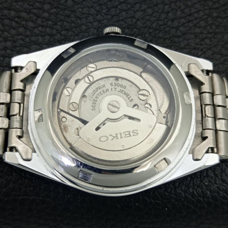 May include: A silver-toned Seiko watch with a transparent back, showcasing the intricate inner workings of the mechanism. The watch face displays the text "JAPAN SEVENTEEN 17 JEWELS" and "SEIKO". The watch band is made of silver metal links.
