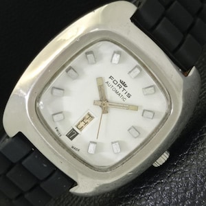 May include: A Fortis Automatic wristwatch with a silver-toned square case, a white dial featuring silver hour markers, and a black rubber strap. The dial displays the brand name, a day-date window at the three o'clock position, and Swiss Made text.
