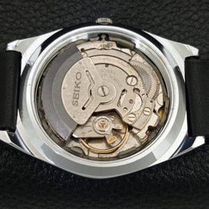 May include: A close-up of the back of a Seiko watch, revealing the intricate mechanical movement. The watch has a silver-colored case and a black leather strap. The movement is labeled "SEIKO" and "17 JEWELS".