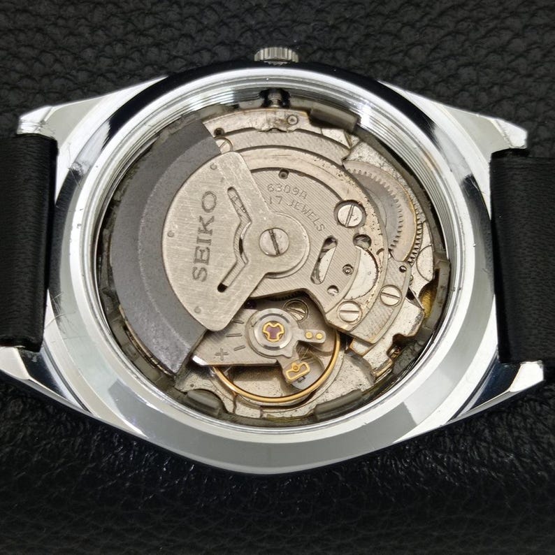 May include: A close-up of the back of a Seiko watch, revealing the intricate mechanical movement. The watch has a silver-colored case and a black leather strap. The movement is labeled "SEIKO" and "17 JEWELS".