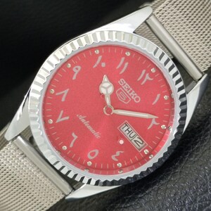 May include: A silver-tone Seiko 5 automatic wristwatch featuring a vibrant red dial with Eastern Arabic numerals. The watch has a fluted bezel, luminous hands, and a day-date window at the three o'clock position, paired with a silver mesh metal strap.
