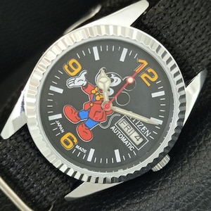 Könnte beinhalten: Eine automatische Citizen-Armbanduhr mit schwarzem Zifferblatt und einem Cartoon-Mickey-Mouse-Design. Die Uhr hat eine silberfarbene Lünette und Zeiger, mit Anzeige von Tag und Datum. Auf dem Zifferblatt steht auch "Japan Made".