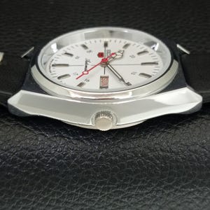 May include: A close-up of a vintage Seiko watch with a white face, black case, and black leather band. The watch features a day and date display, red second hand, and silver-tone hour markers. The watch is on a black textured surface.
