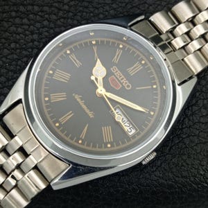 May include: A silver-toned Seiko 5 automatic watch with a dark gray face. The watch features gold-colored hands and Roman numeral hour markers. The watch band is made of silver-toned metal links. The day and date are displayed at the 3 o'clock position.