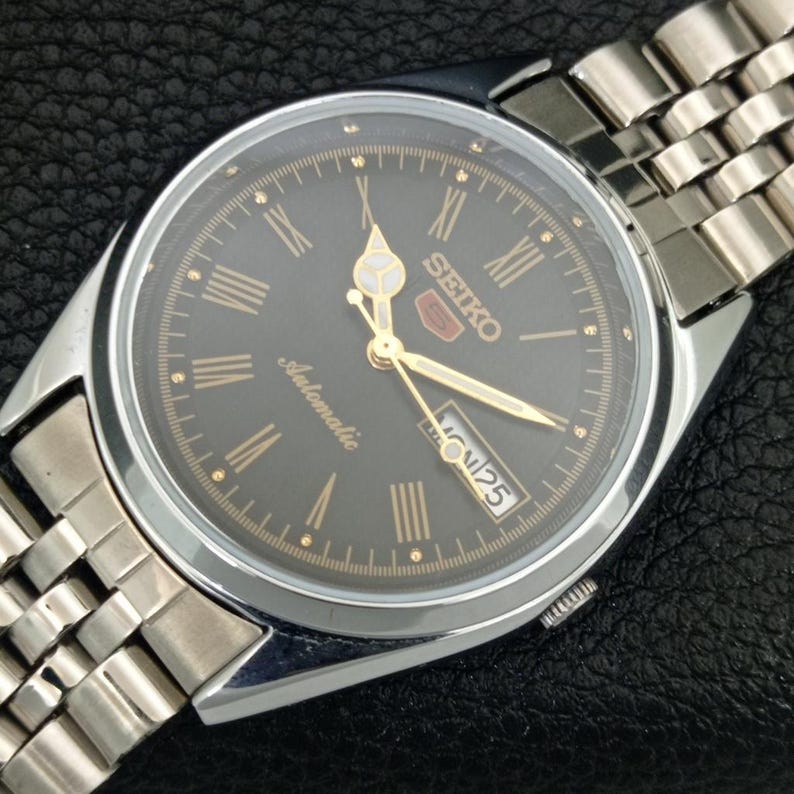 May include: A silver-toned Seiko 5 automatic watch with a dark gray face. The watch features gold-colored hands and Roman numeral hour markers. The watch band is made of silver-toned metal links. The day and date are displayed at the 3 o'clock position.