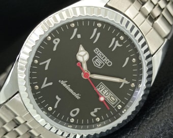 May include: A stainless steel Seiko 5 automatic watch with a black dial and Arabic numerals. The watch features a silver bezel, a silver band, and red second hand. The day of the week is displayed at the 3 o'clock position.