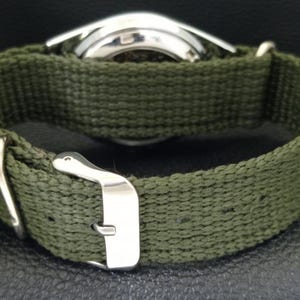 Puede incluir: Primer plano de un reloj de pulsera con una correa de lona verde oliva. La caja del reloj es plateada y la correa tiene una hebilla plateada. El reloj se ve desde atr&aacute;s, mostrando la textura de la correa y el dise&ntilde;o del reloj.
