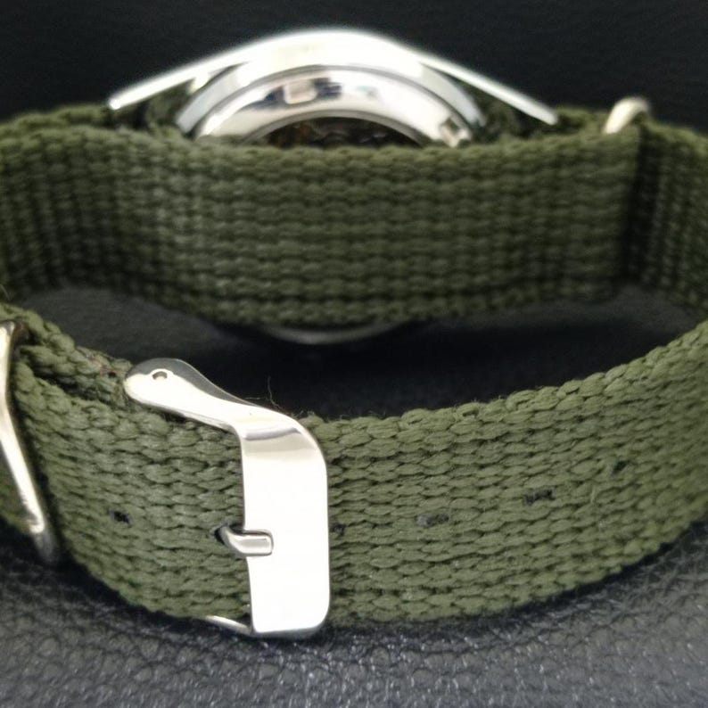Puede incluir: Primer plano de un reloj de pulsera con una correa de lona verde oliva. La caja del reloj es plateada y la correa tiene una hebilla plateada. El reloj se ve desde atr&aacute;s, mostrando la textura de la correa y el dise&ntilde;o del reloj.