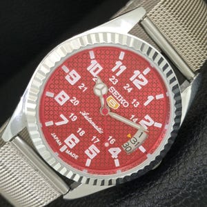 May include: A Seiko 5 automatic wristwatch with a red dial featuring a repeating geometric pattern, white Arabic numerals for 12-hour and 24-hour time, a date window, silver-tone hands, a fluted bezel, and a silver-tone stainless steel mesh strap.