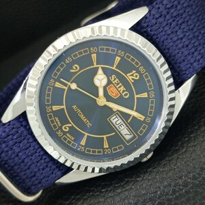 May include: A Seiko 5 automatic wristwatch with a dark blue dial, gold-tone numerals, and hands. The watch features a fluted silver-tone bezel, a day-date window at the three o'clock position, and a textured dark blue fabric strap.