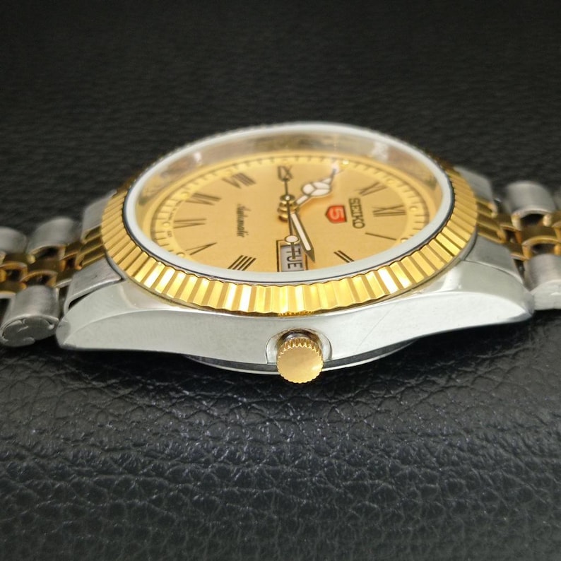 Peut inclure: Une montre automatique Seiko 5 dor&eacute;e avec une lunette dor&eacute;e et un cadran dor&eacute; avec des chiffres romains. La montre a un bracelet argent et or et une couronne dor&eacute;e. Le jour de la semaine est affich&eacute; &agrave; 3 heures.