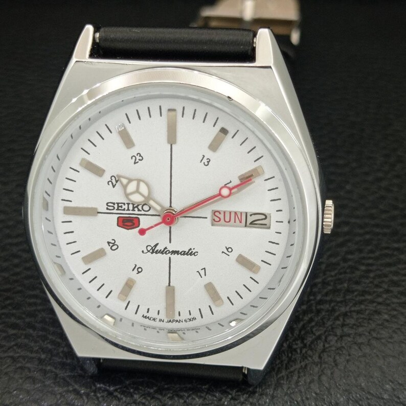 May include: A silver Seiko automatic wristwatch with a white face. The watch features black hour markers, a day and date display, and red accents on the hands. The watch has a black leather band and the text "SEIKO" and "Automatic" are visible on the face.