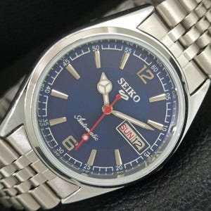 May include: A Seiko 5 automatic wristwatch with a round blue dial, silver-tone hour markers, and a stainless steel link bracelet. The dial features the text Seiko, 5, and Automatic, with a day-date window at the three o'clock position.