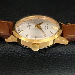 K&ouml;nnte beinhalten: Eine goldfarbene Armbanduhr mit rundem silbernem Zifferblatt, goldenen Zeigern und goldenen Stundenmarkierungen. Das Zifferblatt tr&auml;gt die Aufschrift Montine of Switzerland, 17 Jewels und Incabloc. Die Uhr hat ein braunes Lederarmband mit sichtbaren N&auml;hten.