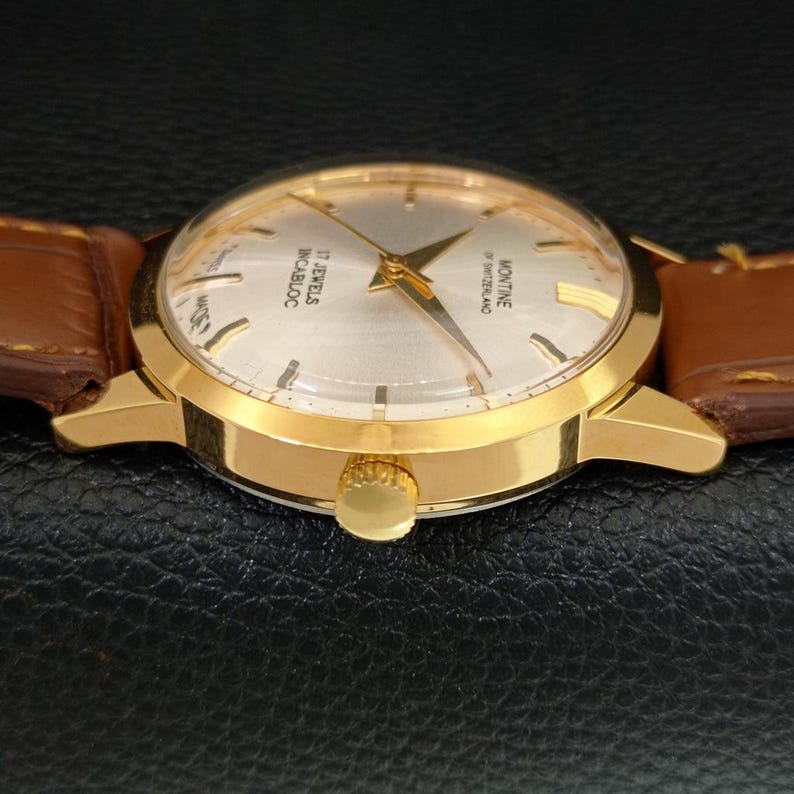 K&ouml;nnte beinhalten: Eine goldfarbene Armbanduhr mit rundem silbernem Zifferblatt, goldenen Zeigern und goldenen Stundenmarkierungen. Das Zifferblatt tr&auml;gt die Aufschrift Montine of Switzerland, 17 Jewels und Incabloc. Die Uhr hat ein braunes Lederarmband mit sichtbaren N&auml;hten.