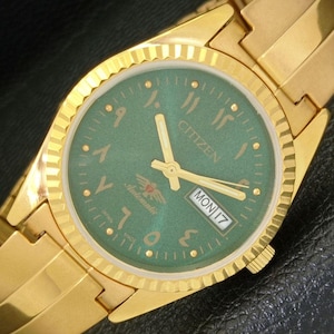 May include: A gold-toned Citizen wristwatch with a green face and Arabic numerals. The watch features gold hands, a day-date display, and the word "Automatic". The watch band is also gold-toned.