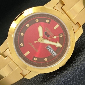 Puede incluir: Reloj automático Seiko 5 dorado con esfera roja. El reloj presenta agujas y marcadores dorados, una pantalla de día y fecha y una pulsera de eslabones dorados. La esfera del reloj tiene la palabra "Automatic" y el logotipo de Seiko.
