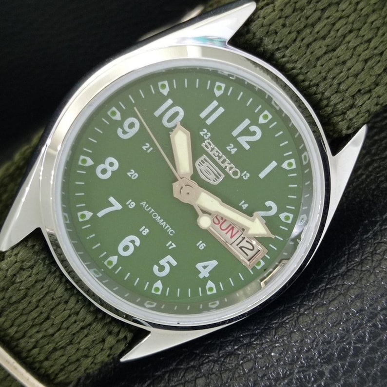 Puede incluir: Primer plano de un reloj autom&aacute;tico Seiko con esfera verde y caja plateada. La esfera muestra el d&iacute;a y la fecha, con n&uacute;meros y agujas blancos. El reloj tiene una correa de tela verde.