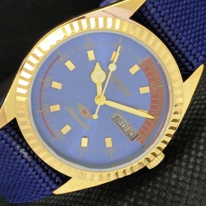 May include: A gold-coloured wristwatch with a blue face and a blue fabric strap. The watch face displays gold hands, markers, and the day of the week. The watch has the word "Citizen" and "Automatic" printed on the face.