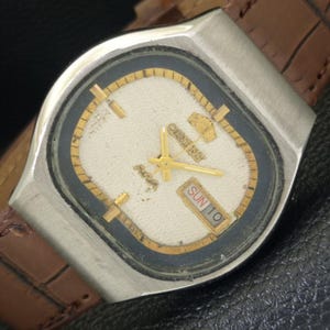 May include: A vintage Orient watch with a silver-tone case and a brown leather strap. The watch face is off-white with gold-tone hands and markers, and a day/date display. The watch is set to "SUN 10".