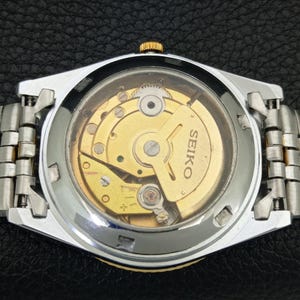 Peut inclure: Gros plan du dos d'une montre Seiko, r&eacute;v&eacute;lant le mouvement m&eacute;canique dor&eacute; complexe visible &agrave; travers un verre transparent. La montre a un bracelet en m&eacute;tal argent&eacute; et dor&eacute;.