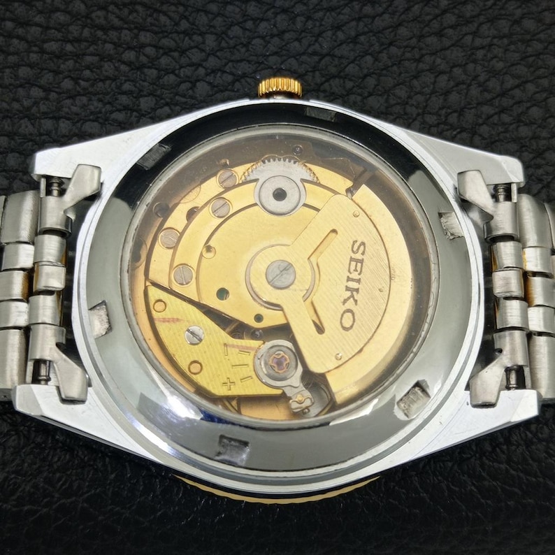 Peut inclure: Gros plan du dos d'une montre Seiko, r&eacute;v&eacute;lant le mouvement m&eacute;canique dor&eacute; complexe visible &agrave; travers un verre transparent. La montre a un bracelet en m&eacute;tal argent&eacute; et dor&eacute;.