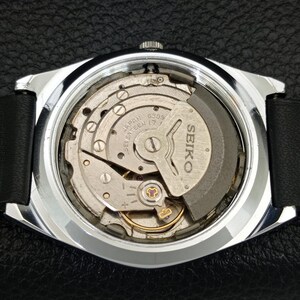 May include: A close-up of the back of a Seiko watch, showcasing the intricate internal mechanism. The watch has a silver-colored case and a black leather strap. The internal components are silver and gold, with the Seiko logo visible.