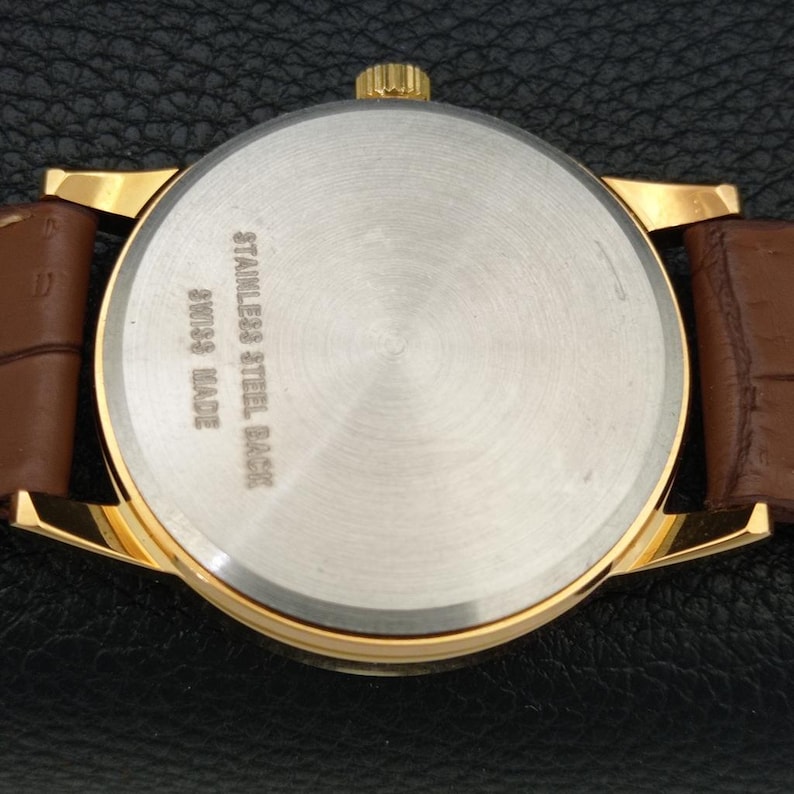 K&ouml;nnte beinhalten: Eine goldfarbene Armbanduhr mit einem braunen Lederarmband mit strukturiertem Muster. Der runde Geh&auml;useboden besteht aus Edelstahl und ist mit dem Text STAINLESS STEEL BACK und SWISS MADE sowie einer kleinen goldfarbenen Krone versehen.