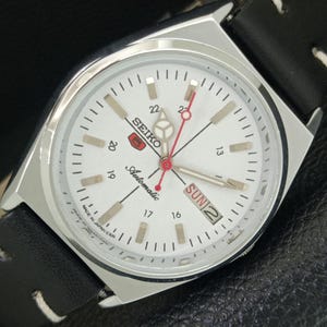 May include: A silver Seiko automatic watch with a white face, black leather band, and day/date display. The watch features a red second hand and luminous markers. The watch face displays the text "SEIKO Automatic" and "SUN 2".