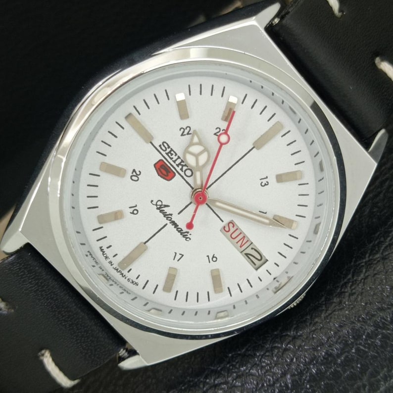 May include: A silver Seiko automatic watch with a white face, black leather band, and day/date display. The watch features a red second hand and luminous markers. The watch face displays the text "SEIKO Automatic" and "SUN 2".