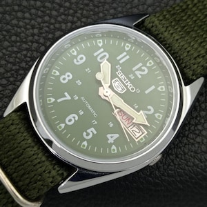 Puede incluir: Un reloj autom&aacute;tico Seiko con esfera verde y correa de tela verde. El reloj tiene una caja plateada y presenta n&uacute;meros y agujas blancas. El d&iacute;a y la fecha se muestran a las 3 en punto. El reloj est&aacute; sobre una superficie negra.