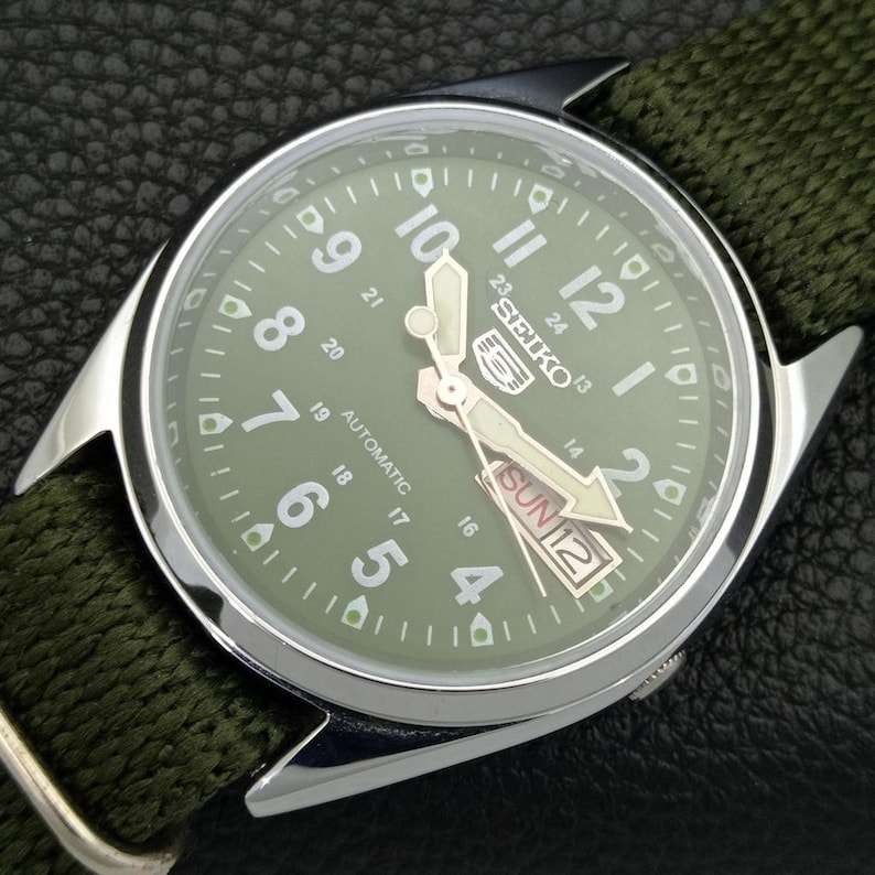 Puede incluir: Un reloj autom&aacute;tico Seiko con esfera verde y correa de tela verde. El reloj tiene una caja plateada y presenta n&uacute;meros y agujas blancas. El d&iacute;a y la fecha se muestran a las 3 en punto. El reloj est&aacute; sobre una superficie negra.