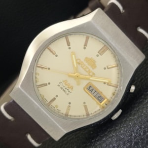 May include: A vintage Orient AAA Crystal 2 Jewels watch with a cream-colored face, gold hands and markers, and a day-date display. The watch has a silver-tone case and a brown leather strap.