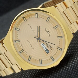 Puede incluir: Un reloj de pulsera dorado con una esfera blanca y marcas negras. El reloj tiene una ventana de día y fecha, y el texto "FORTIS EDEN ROC AUTOMATIC" está impreso en la esfera. El reloj está en una banda de metal dorado.