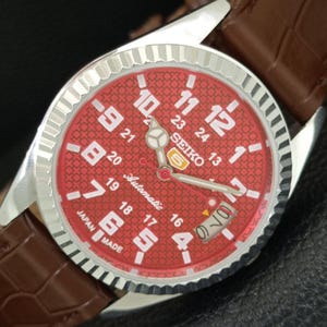 May include: A Seiko 5 automatic wristwatch with a silver-tone fluted bezel and a textured red dial. The dial features large white Arabic numerals, a 24-hour scale, luminous hands, and a date window. It is attached to a brown leather strap.