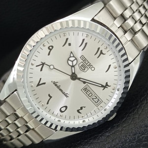Könnte beinhalten: Eine silberne Seiko 5 Automatik-Armbanduhr mit arabischen Ziffern auf dem Zifferblatt. Die Uhr hat ein silbernes Armband und eine Datumsanzeige mit "MIT 23". Die Zeiger der Uhr sind schwarz, und das Zifferblatt ist silberfarben.