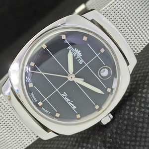 May include: A silver Fortis Trueline wristwatch with a black face and a silver mesh strap. The watch face has a grid pattern and the words "Fortis Trueline" printed on it. The watch also has a small date window at the 6 o'clock position.
