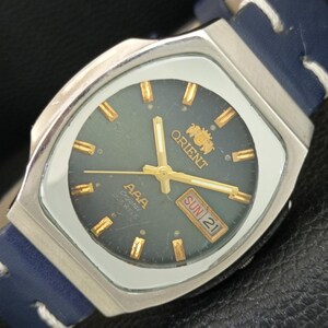 May include: An Orient AAA Crystal watch with a dark gray dial, gold-tone hour markers, and gold hands. The watch features a day-date window at the three o'clock position displaying SUN 21, housed in a silver-tone case with a blue leather strap.