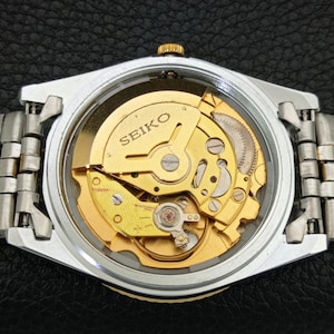 Peut inclure: Gros plan d'une montre automatique Seiko, r&eacute;v&eacute;lant le m&eacute;canisme interne dor&eacute; &agrave; travers un dos transparent. La montre a un bo&icirc;tier argent&eacute; et un bracelet en m&eacute;tal. Le mot "SEIKO" est visible sur le mouvement.
