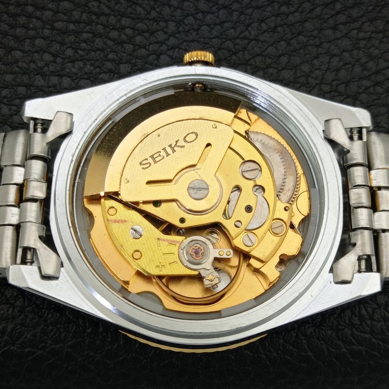 Peut inclure: Gros plan d'une montre automatique Seiko, r&eacute;v&eacute;lant le m&eacute;canisme interne dor&eacute; &agrave; travers un dos transparent. La montre a un bo&icirc;tier argent&eacute; et un bracelet en m&eacute;tal. Le mot "SEIKO" est visible sur le mouvement.